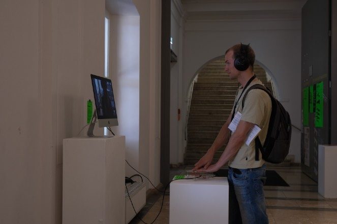 A visitor wearing headphones stands at the Being, Between installation next to a screen on a plinth.